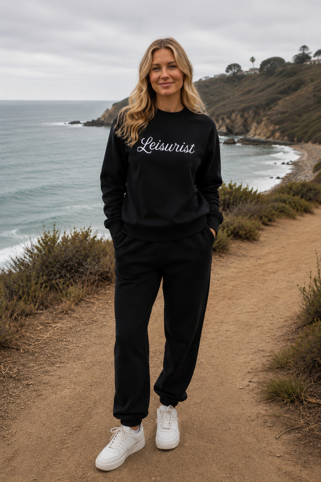 Woman in a black embroidered Leisurist script sweatshirt standing on a coastal path with ocean and cliffs in the background.